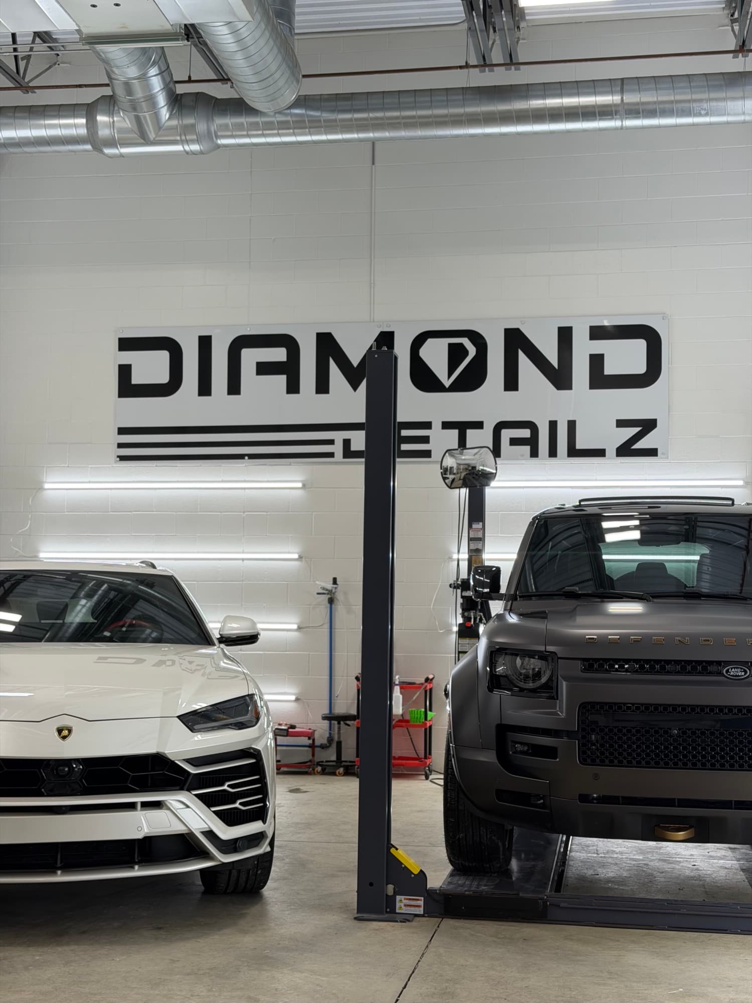 White Lamborghini Urus and matte-black Land Rover Defender side-by-side in the Diamond Detailz install bay under the brand logo wall