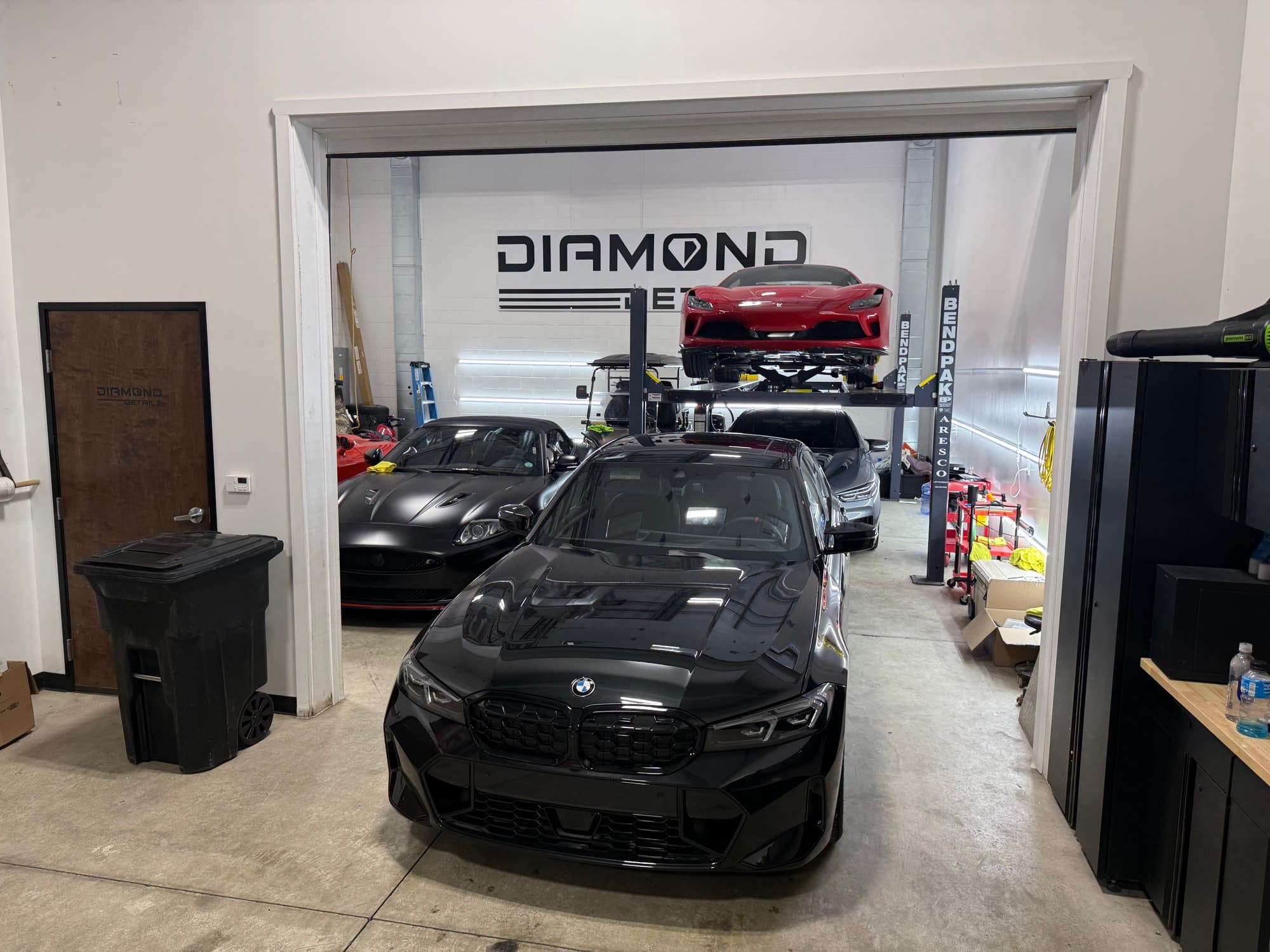 Black BMW M3 G80 framed by the Diamond Detailz shop doorway with the brand logo wall behind it