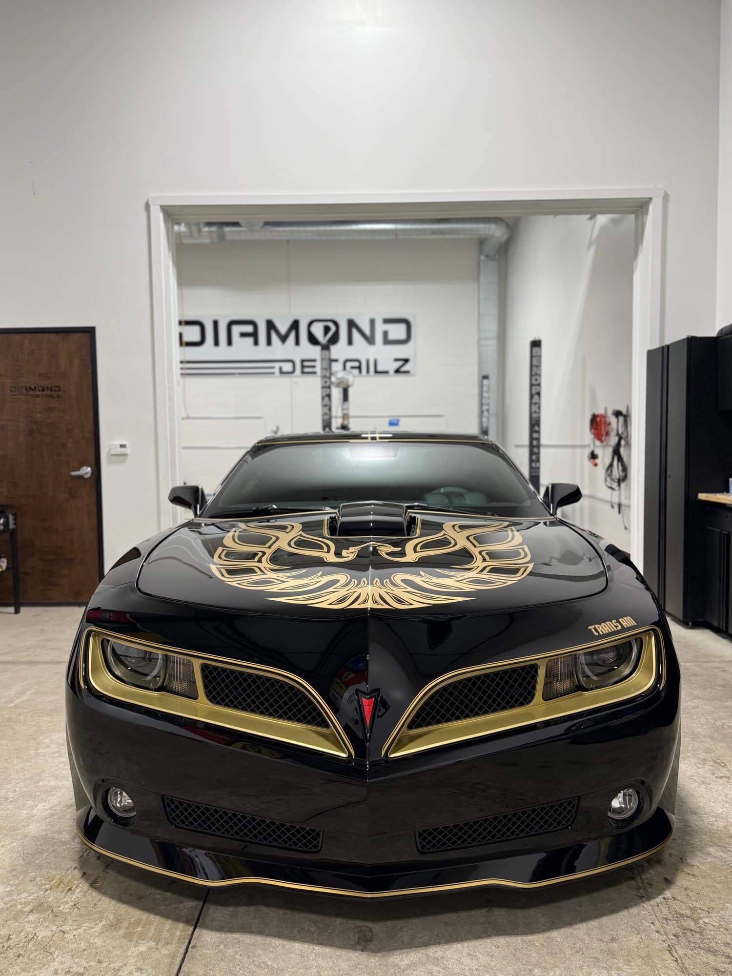 Black 2nd-gen Pontiac Trans Am head-on with the gold Firebird hood emblem framed under the Diamond Detailz logo wall — the customer car corrected in the slider below.